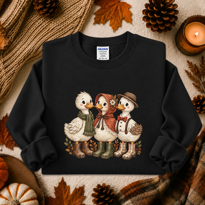 A black crewneck sweatshirt featuring a graphic of three cute ducks dressed in autumn clothing, styled in a cozy fall flat-lay with pinecones, leaves, a candle, and a knit blanket.