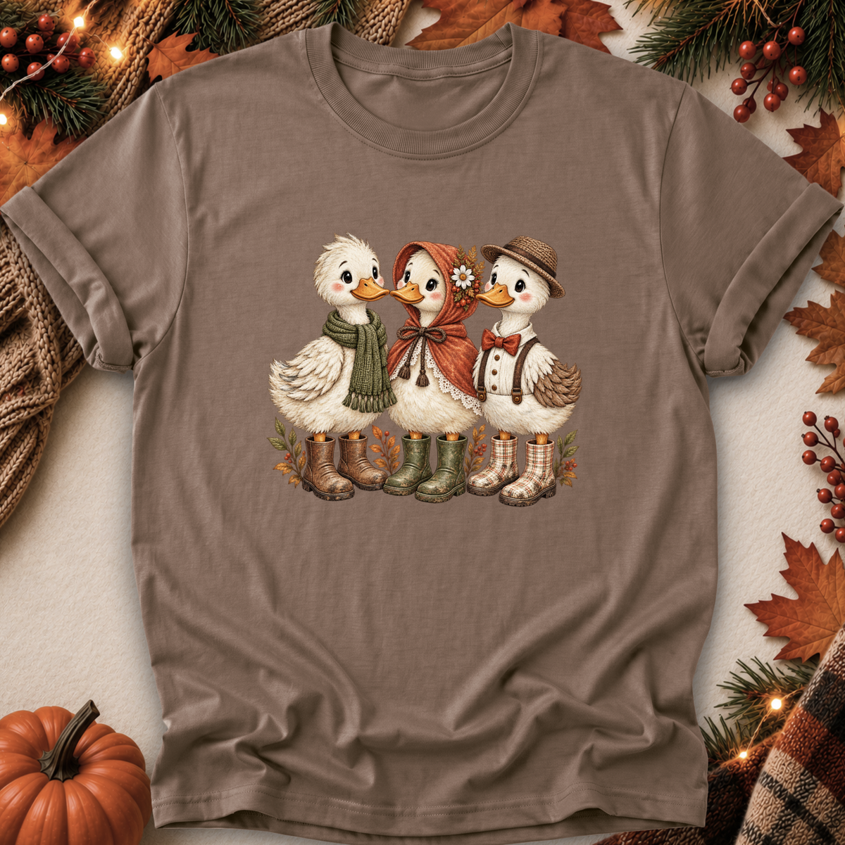A taupe brown short-sleeve crewneck t-shirt featuring a graphic of three cute cartoon ducks dressed in autumn clothing and boots, laid flat on a neutral background with seasonal props like a pumpkin, pine branches, and fall leaves.