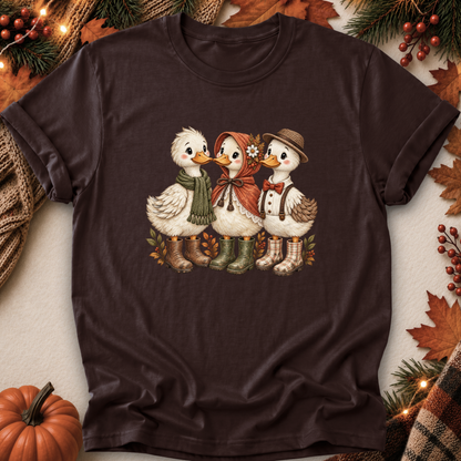 A dark brown short-sleeve t-shirt laid flat on a light beige surface, surrounded by autumn-themed props including a pumpkin, pine branches, leaves, and string lights. The shirt features a graphic print of three cute cartoon ducks dressed in fall clothing.