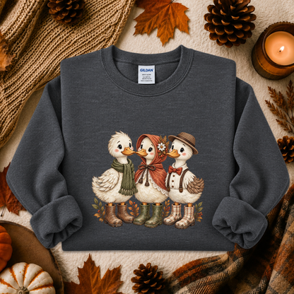 A dark gray crewneck sweatshirt featuring a graphic of three cute ducks dressed in autumn clothing, styled in a cozy fall flat-lay with pinecones, leaves, a candle, and a knit blanket.