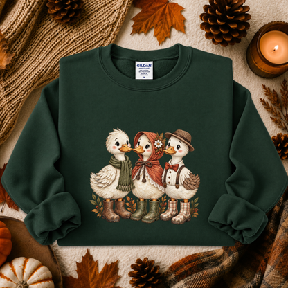 A dark green crewneck sweatshirt featuring a graphic of three cute ducks dressed in autumn clothing, styled in a cozy fall flat-lay with pinecones, a candle, and a knit blanket.