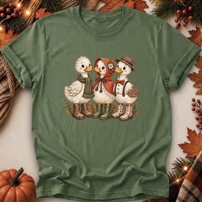 An olive green short-sleeve t-shirt with a graphic of three cute cartoon ducks dressed in autumn clothing and boots, laid flat on a neutral background with seasonal props like a pumpkin, pine branches, and fall leaves.