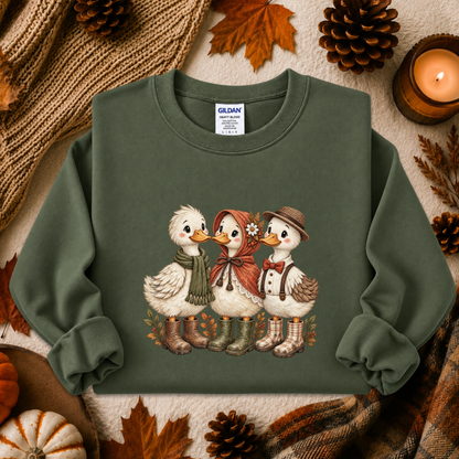An olive green long-sleeve crewneck sweatshirt featuring a graphic of three cute ducks in autumn clothing, styled in a cozy fall flat-lay with pinecones, a candle, and a knit blanket.