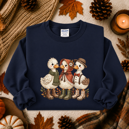 A navy blue crewneck sweatshirt featuring a graphic of three cute cartoon ducks in autumn clothing, styled in a cozy fall flat-lay with pinecones, leaves, a candle, and a knit blanket.