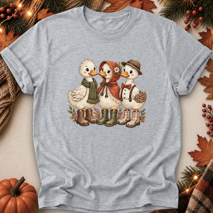 A light heather gray short-sleeve t-shirt featuring a vintage-style graphic of three cute ducks dressed in autumn clothing, surrounded by a cozy fall-themed flat-lay background with pumpkins, pine branches, and leaves.