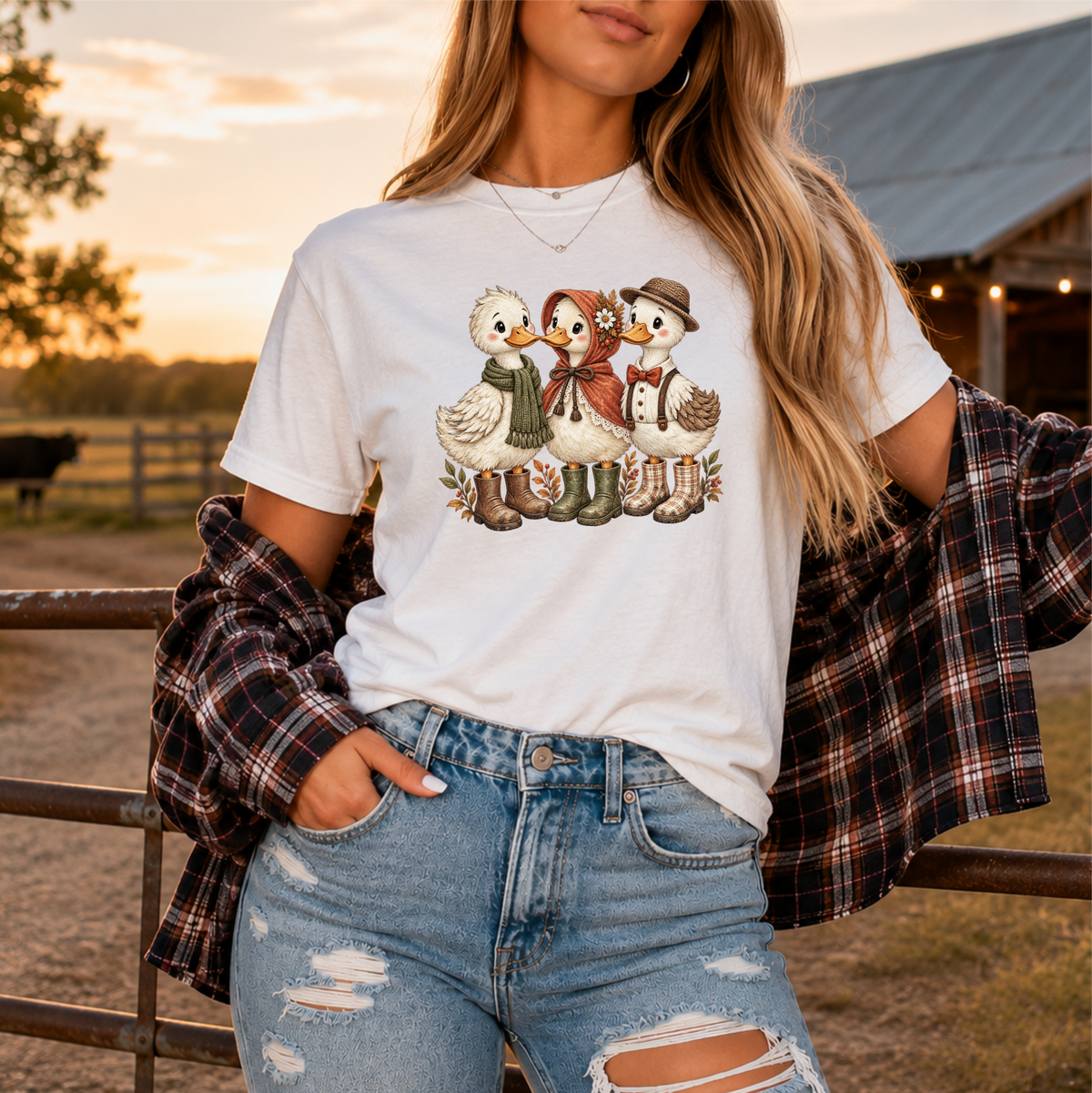 A woman wearing a white graphic t-shirt with a cute cartoon duck family design, styled with a black and red plaid flannel shirt tied around her shoulders and light-wash distressed jeans, set against a warm sunset farm background.