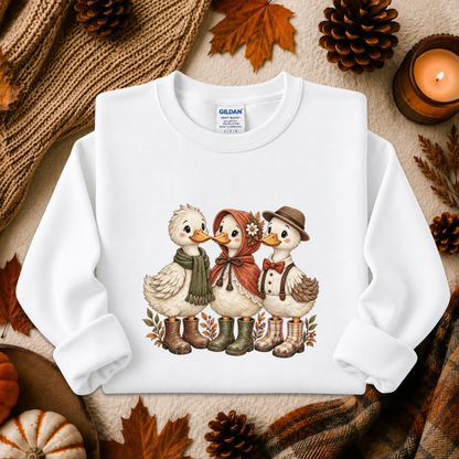 A white long-sleeve sweatshirt laid flat on a cozy autumn-themed background. The sweatshirt features a printed graphic of three cute ducks dressed in fall clothing: one in a green scarf, one in a red hood with flowers, and one in a brown hat and suspenders.
