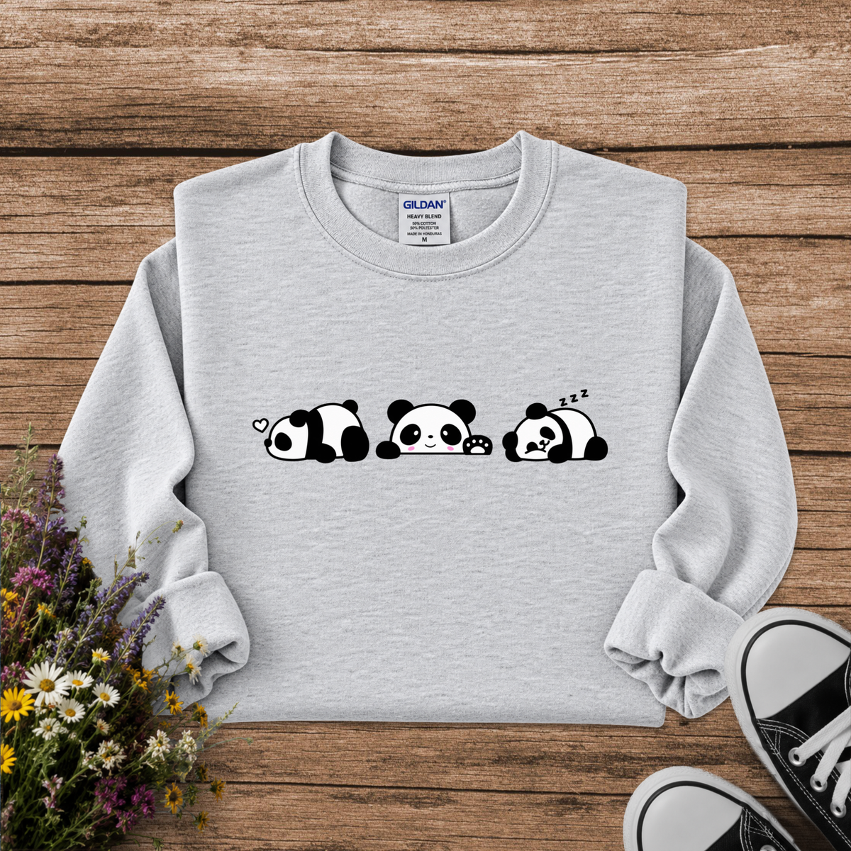 A light gray sweatshirt laid flat on a rustic wooden surface, featuring a black-and-white panda face graphic across the chest. The sweatshirt is styled with a bouquet of wildflowers on the left and black-and-white sneakers at the bottom right.