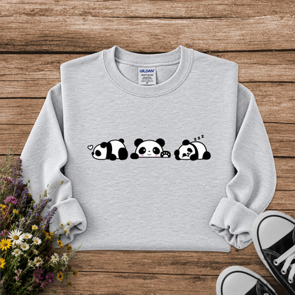 A light gray sweatshirt laid flat on a rustic wooden surface, featuring a black-and-white panda face graphic across the chest. The sweatshirt is styled with a bouquet of wildflowers on the left and black-and-white sneakers at the bottom right.