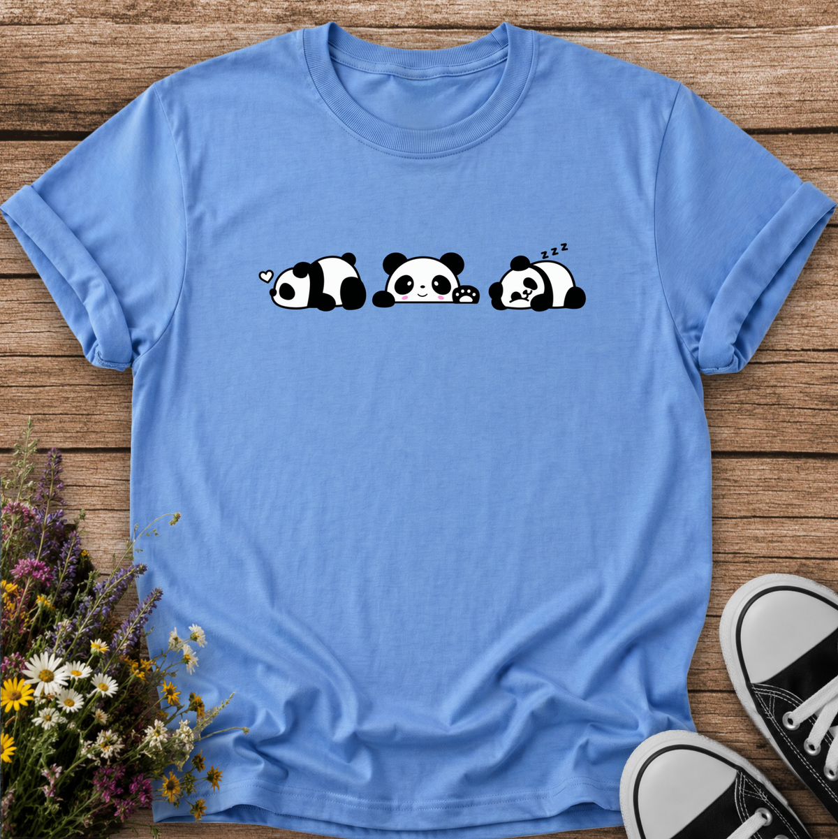 A light blue short-sleeve t-shirt with a cute cartoon panda graphic, laid flat on a rustic wooden background with wildflowers and black-and-white sneakers as props.