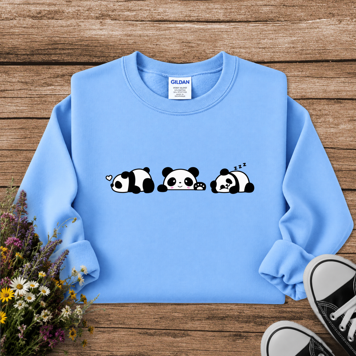 A light blue crewneck sweatshirt with a black and white cartoon panda graphic across the chest, styled in a flat lay on a rustic wooden background with wildflowers and black sneakers.