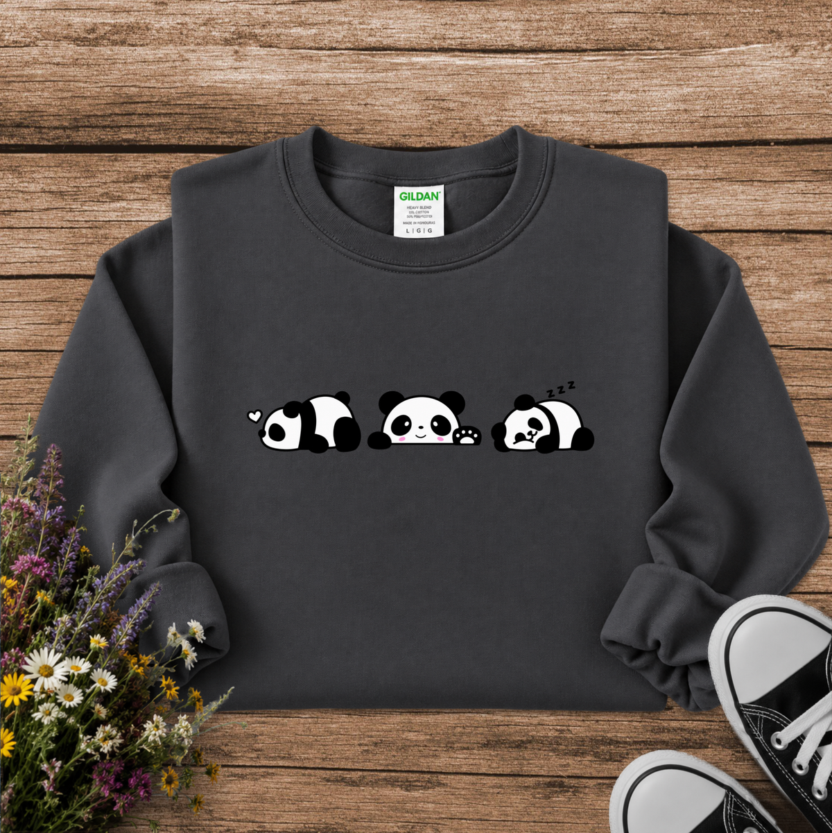 A dark charcoal gray sweatshirt with a cute cartoon panda graphic, laid flat on a rustic wooden background with wildflowers and black canvas sneakers as props.