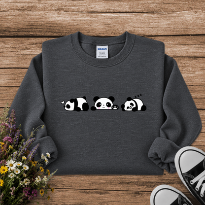 A dark gray crewneck sweatshirt featuring a cute graphic of three sleeping pandas in a row. The sweatshirt is displayed in a flat-lay arrangement on a rustic wooden background, styled with a small bouquet of wildflowers on the left and the corner of a black canvas sneaker on the bottom right.
