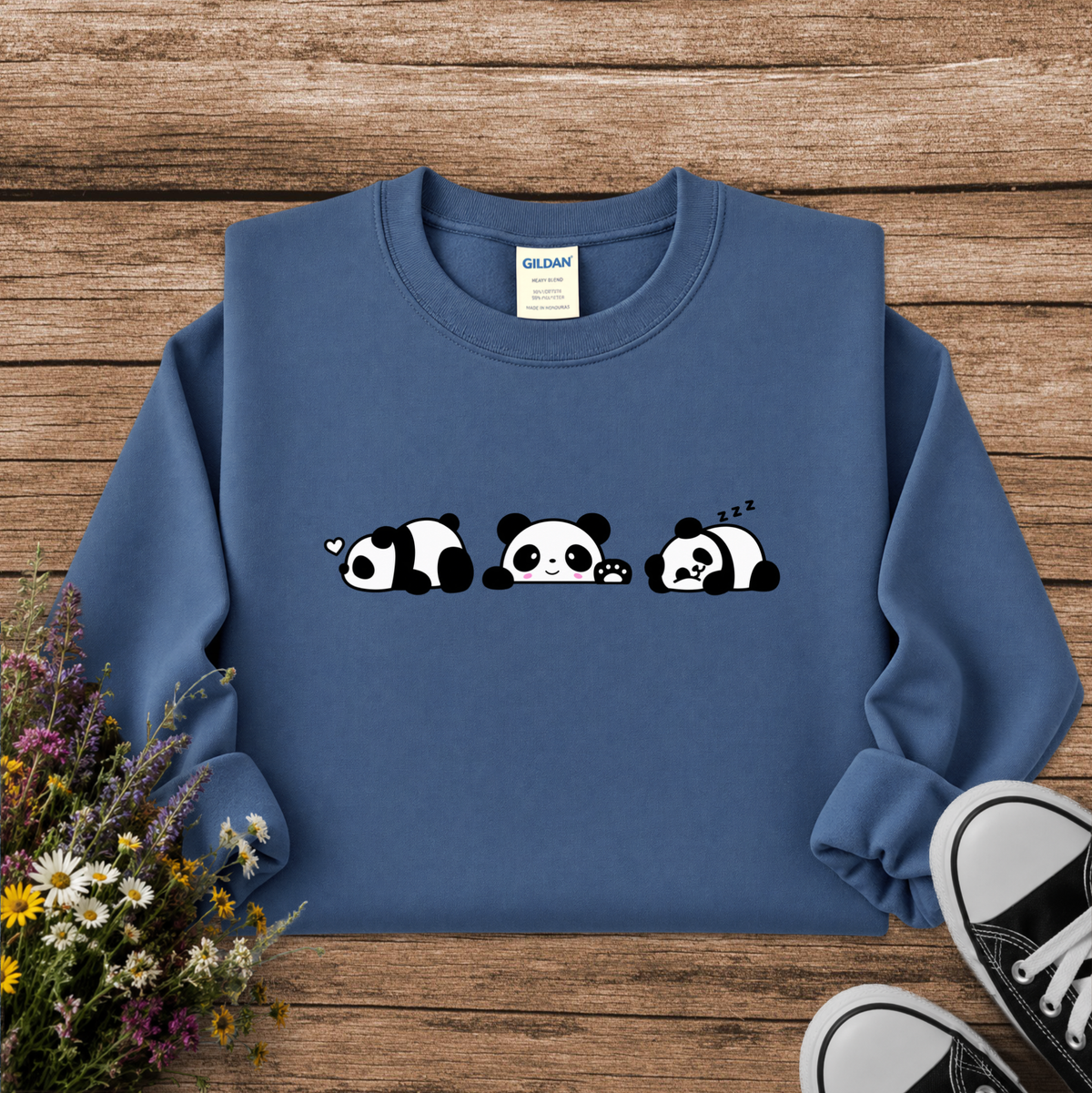 A blue crewneck sweatshirt featuring a cute cartoon panda graphic in three sleeping poses across the chest. The sweatshirt is displayed in a flat-lay arrangement on a rustic wooden background, styled with a small bouquet of wildflowers on the left and black canvas sneakers on the bottom right.