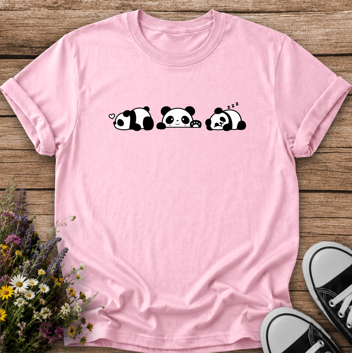 A light pink short-sleeve t-shirt with a black and white cartoon panda graphic, laid flat on a rustic wooden background with flowers and sneakers as props.