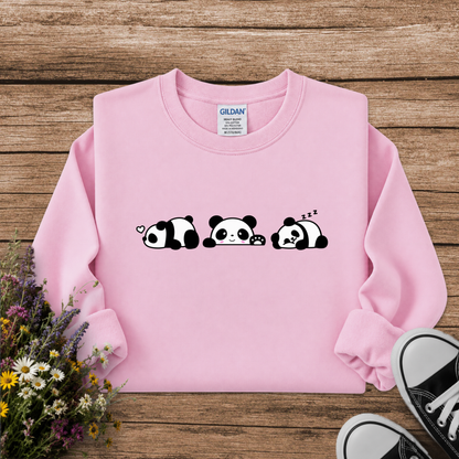 A light pink long-sleeve sweatshirt with a black and white cartoon panda graphic across the chest, styled in a flat lay on a rustic wooden background with flowers and black sneakers.