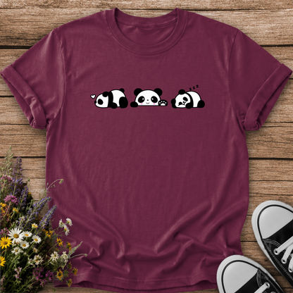 A maroon short-sleeve t-shirt with a black and white cartoon panda graphic, laid flat on a wooden surface with flowers and sneakers as props.