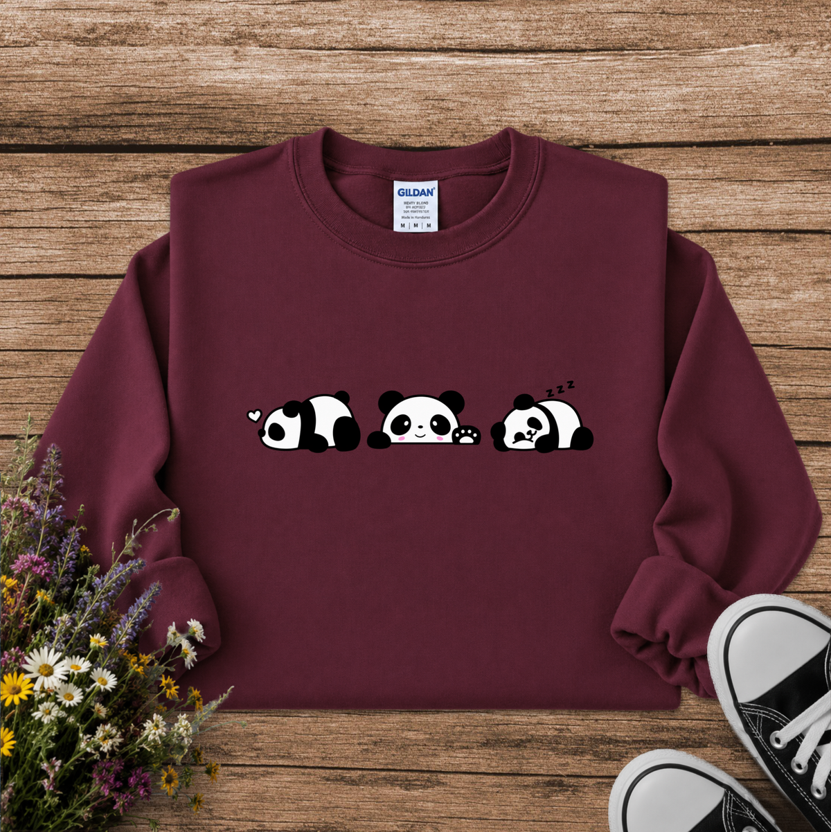 A maroon crewneck sweatshirt featuring a cute cartoon panda graphic across the chest, styled in a flat lay on a rustic wooden background with flowers and black sneakers.