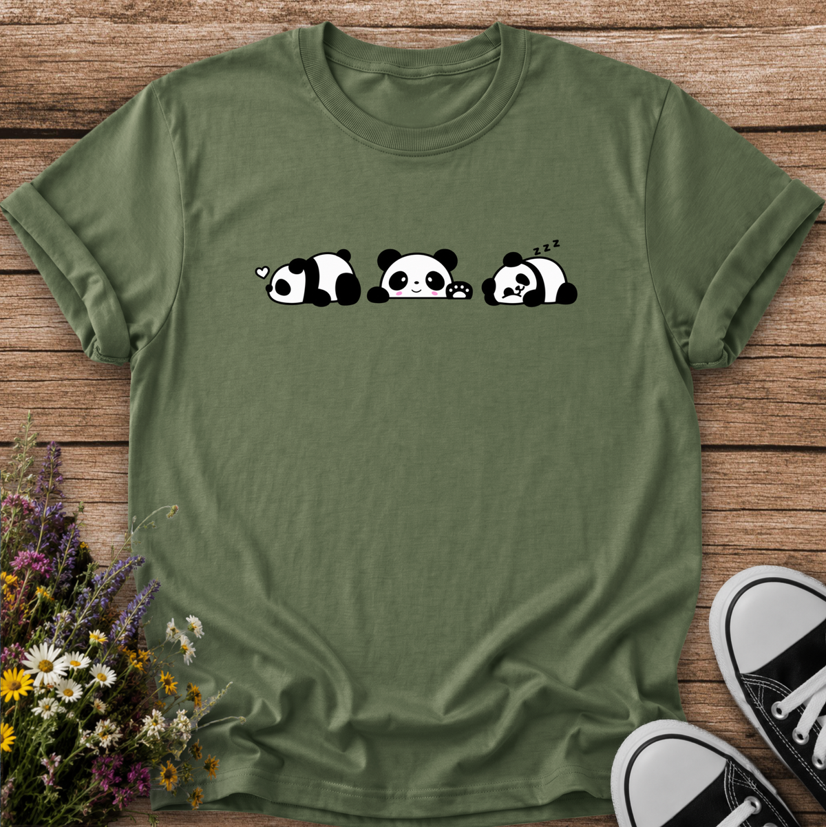 An olive green short-sleeve t-shirt with a graphic of three cute sleeping pandas, laid flat on a rustic wooden background with wildflowers and black-and-white sneakers as props.