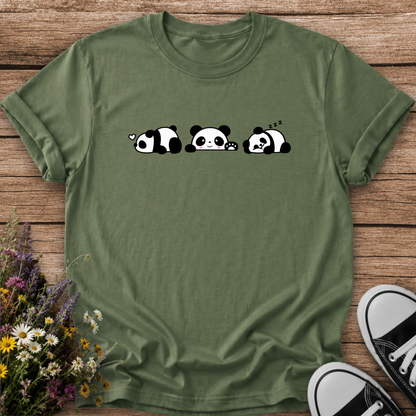 An olive green short-sleeve t-shirt with a graphic of three cute sleeping pandas, laid flat on a rustic wooden background with wildflowers and black-and-white sneakers as props.