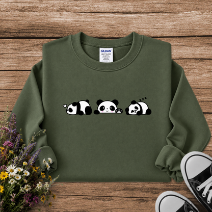 An olive green crewneck sweatshirt featuring a cute graphic of three sleeping pandas on the chest, styled in a flat-lay on a rustic wooden background with wildflowers and black sneakers.