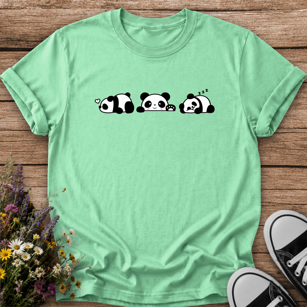 A mint green short-sleeve t-shirt with a black and white cartoon panda graphic, laid flat on a wooden surface with flowers and black-and-white sneakers as props.
