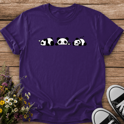 A purple short-sleeve t-shirt with a cartoon panda graphic, laid flat on a wooden surface with flowers and sneakers as props.
