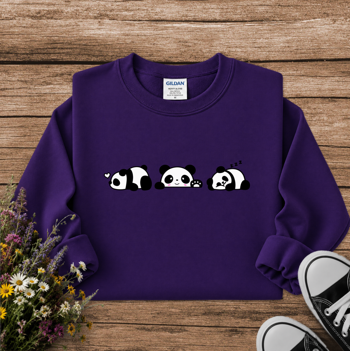 A purple crewneck sweatshirt featuring a cute graphic of three sleeping pandas in black and white. The sweatshirt is laid flat on a rustic wooden background, styled with a small bouquet of wildflowers on the left and black canvas sneakers on the right.
