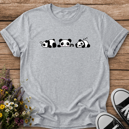 A light heather gray short-sleeve t-shirt with a black and white cartoon panda graphic, laid flat on a rustic wooden background with flowers and black-and-white sneakers as props.