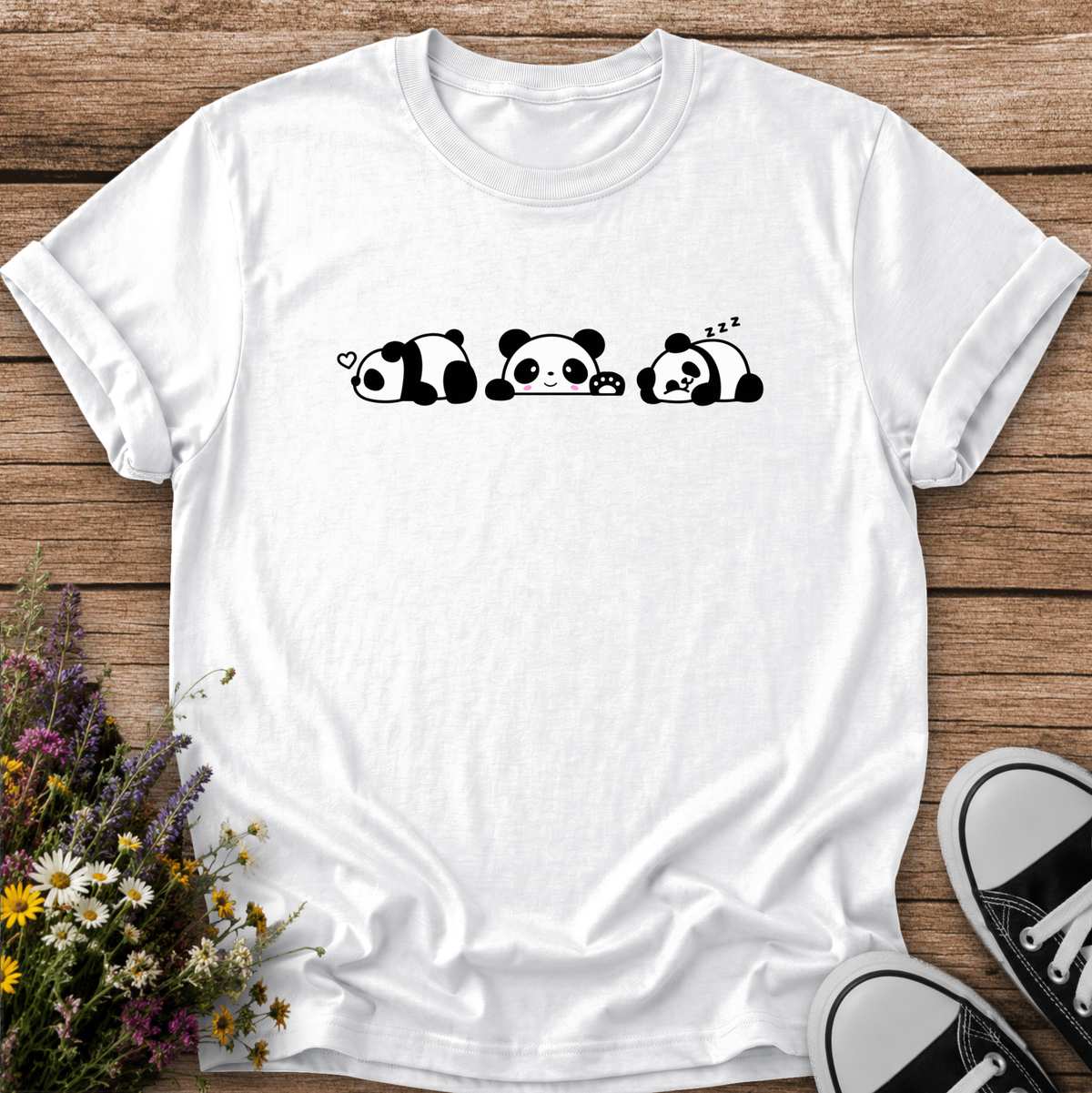 A white short-sleeve crewneck t-shirt featuring a black line-art graphic of three sleeping pandas, laid flat on a rustic wooden background with flowers and black-and-white sneakers as props.
