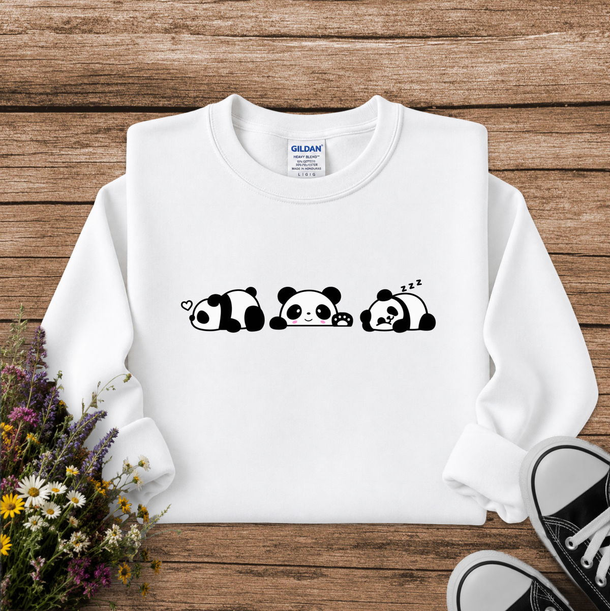 A white long-sleeve crewneck sweatshirt with a black cartoon panda graphic across the chest, laid flat on a rustic wooden background with wildflowers and black sneakers as props. The Gildan brand tag is visible inside the collar.