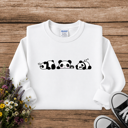A white long-sleeve crewneck sweatshirt with a black cartoon panda graphic across the chest, laid flat on a rustic wooden background with wildflowers and black sneakers as props. The Gildan brand tag is visible inside the collar.