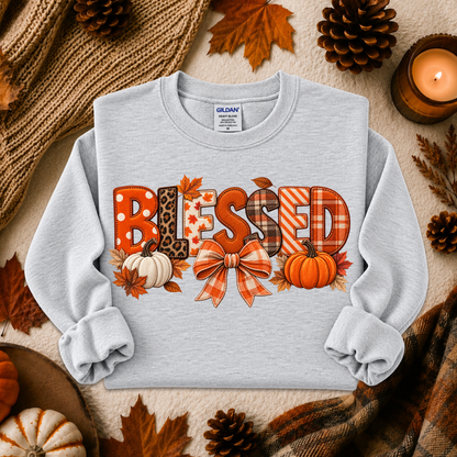 A light gray long-sleeve crewneck sweatshirt with a 'Blessed' autumn-themed graphic, styled in a flat lay with fall leaves, pinecones, a candle, and a knitted scarf.