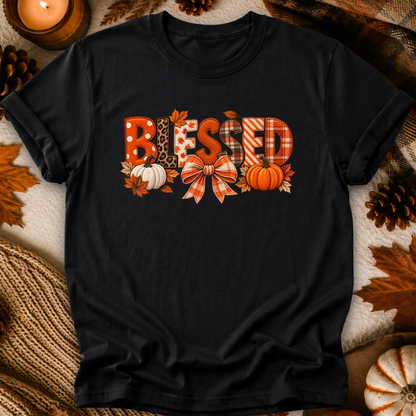 A black short-sleeve t-shirt with a 'Blessed' autumn graphic, laid flat on a cozy background of faux fur, pinecones, and a lit candle. The graphic features the word 'Blessed' in various fall-themed patterns (plaid, leopard print, polka dots) decorated with pumpkins, leaves, and a bow.