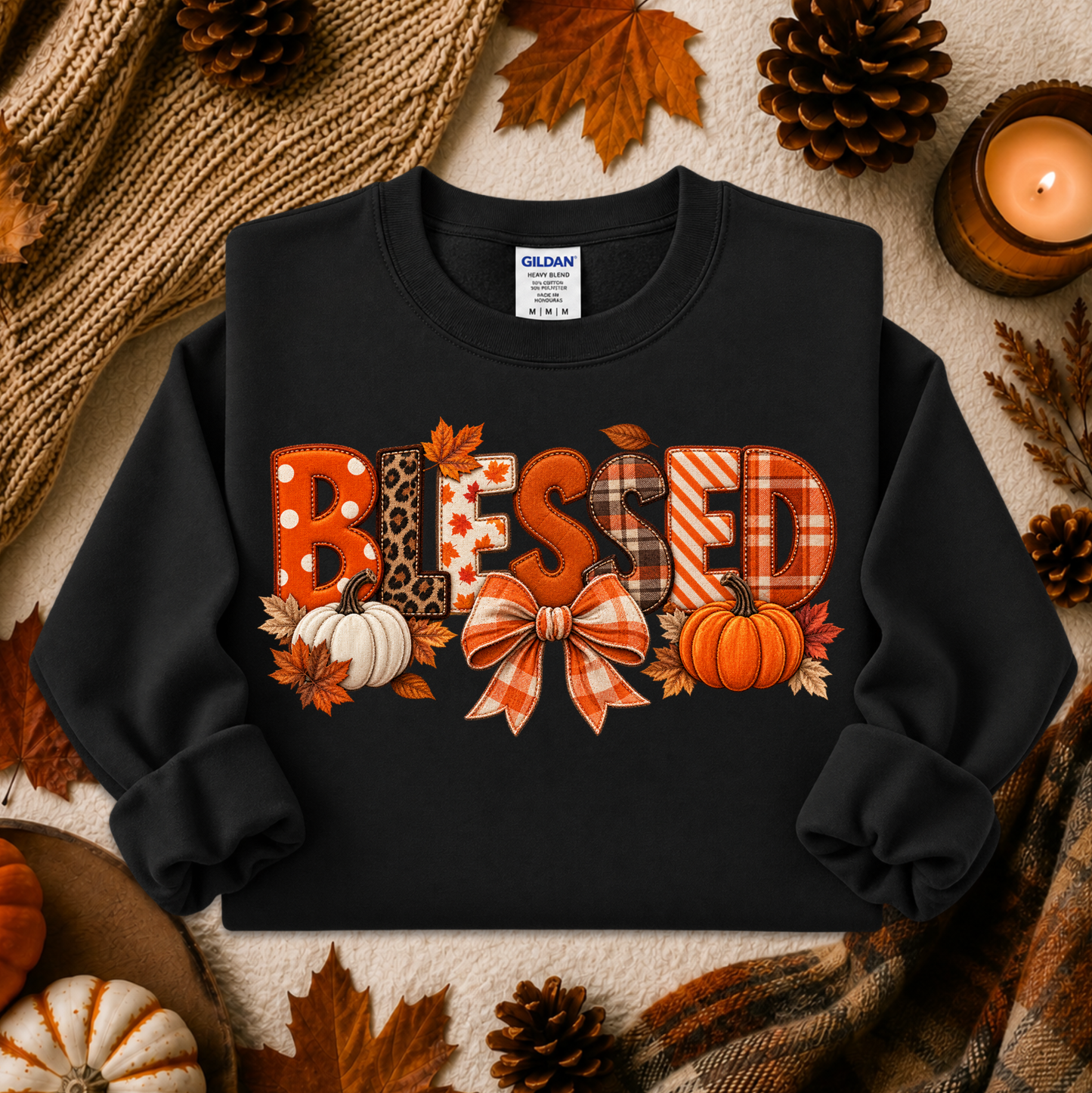 A black crewneck sweatshirt with a 'BLESSED' graphic made of autumn-themed patterned letters, featuring pumpkins, leaves, and a bow, styled in a cozy fall flat-lay with pinecones, a candle, and a knit blanket.