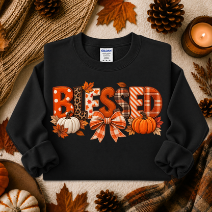 A black crewneck sweatshirt with a 'BLESSED' graphic made of autumn-themed patterned letters, featuring pumpkins, leaves, and a bow, styled in a cozy fall flat-lay with pinecones, a candle, and a knit blanket.
