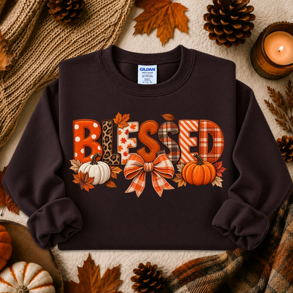 A dark brown crewneck sweatshirt featuring a 'BLESSED' autumn-themed graphic with pumpkins, leaves, and patterned letters, styled in a cozy flat-lay with fall props.