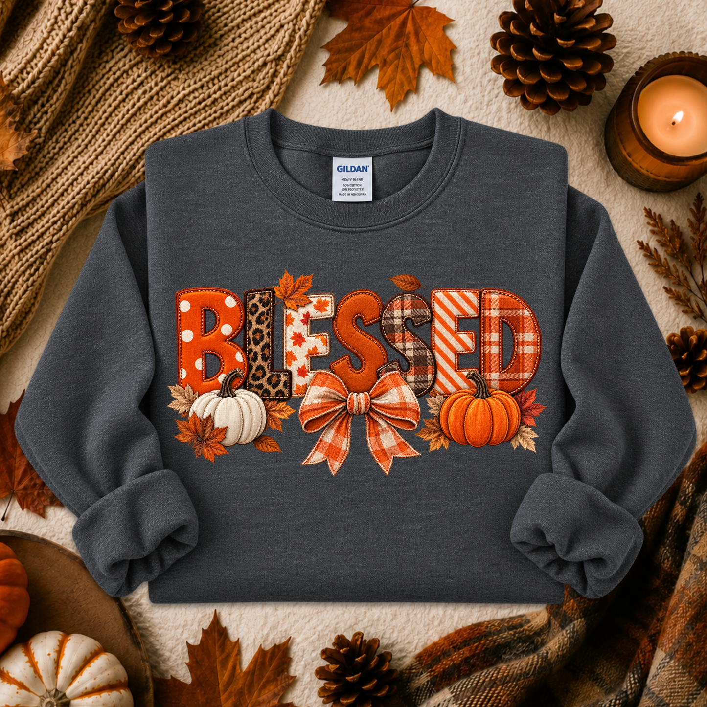 A dark gray crewneck sweatshirt featuring a 'BLESSED' autumn-themed graphic with pumpkins, leaves, and a bow, styled in a flat lay with fall decorations.