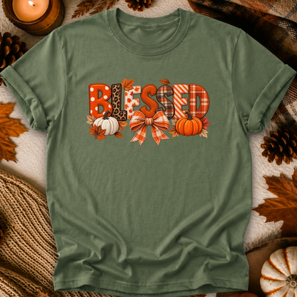 An olive green short-sleeve t-shirt with a 'Blessed' autumn graphic, laid flat on a cozy background of faux fur, pinecones, and a lit candle.