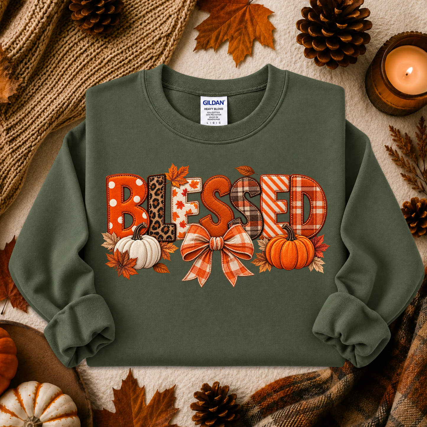 An olive green crewneck sweatshirt with a 'BLESSED' autumn graphic, styled in a cozy flat-lay with pumpkins, pinecones, a candle, and a knitted blanket. The brand Gildan is visible on the inner neck label.
