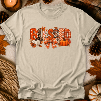 A light beige graphic t-shirt laid flat on a cozy autumn-themed background with a lit candle, pinecones, and leaves. The shirt features a 'BLESSED' graphic with various fall patterns like leopard print, plaid, polka dots, and stripes, decorated with pumpkins and a bow.