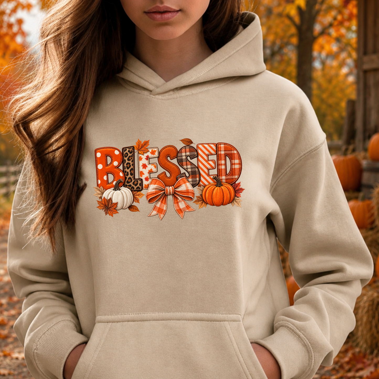 A woman wearing a beige hoodie with a 'Blessed' autumn graphic featuring pumpkins, leaves, and patterned letters, set against an outdoor fall background.