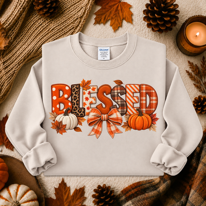 A light beige crewneck sweatshirt with a 'Blessed' autumn graphic, styled in a cozy flat-lay with fall leaves, pinecones, a candle, and a knitted blanket.