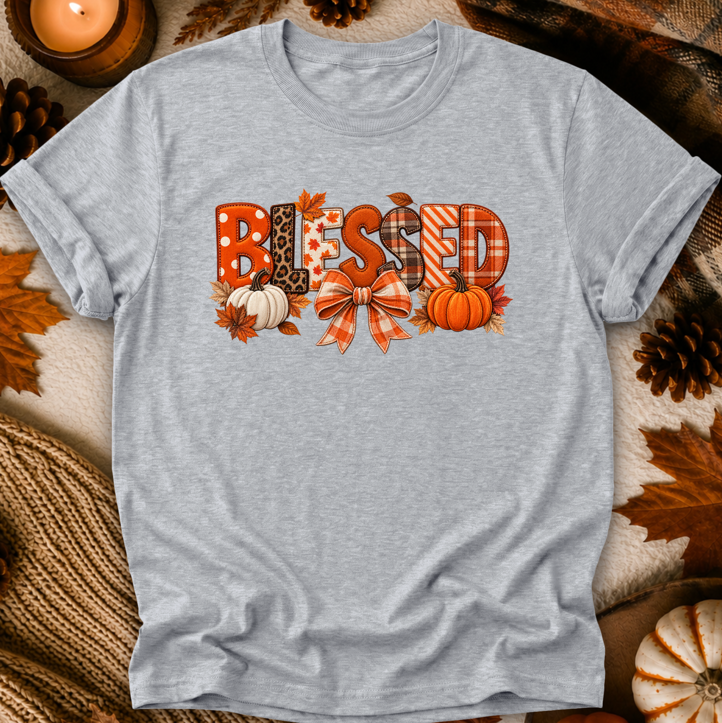 A light heather gray short-sleeve t-shirt with a 'Blessed' autumn graphic, laid flat on a cozy fall-themed background with pinecones, leaves, and a candle.