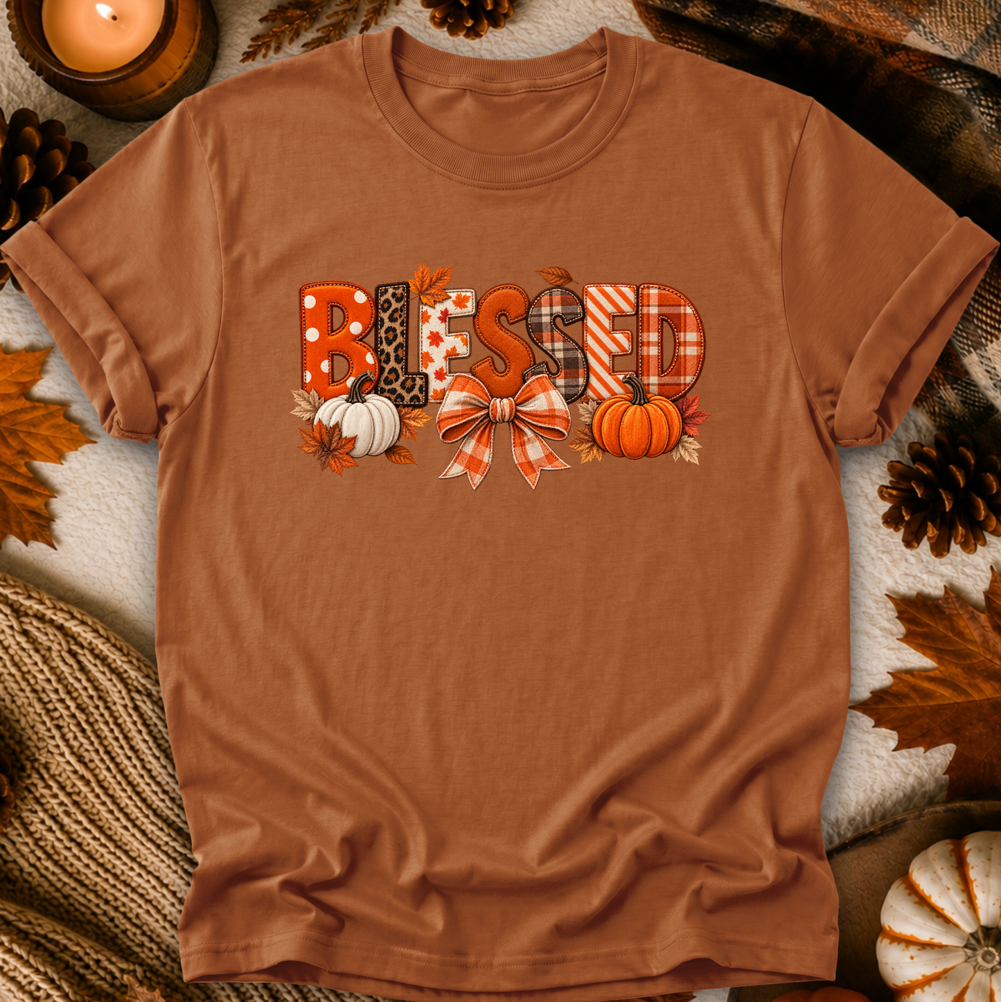 A rust-brown short-sleeve t-shirt laid flat with a decorative 'Blessed' graphic on the chest, surrounded by autumn props including pumpkins, pinecones, leaves, and a candle.
