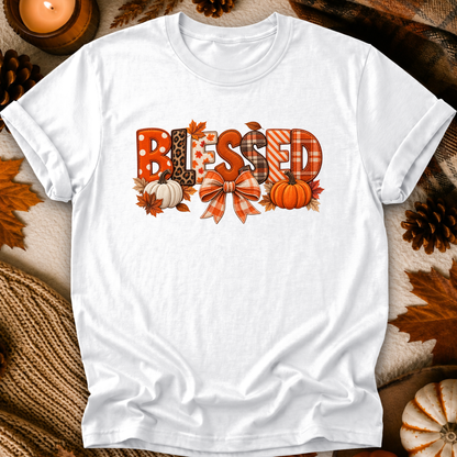 A white short-sleeve crewneck t-shirt laid flat on a cozy autumn-themed background. The shirt features a graphic spelling 'BLESSED' in various fall patterns (plaid, leopard print, stripes, polka dots) decorated with orange pumpkins, leaves, and a bow.