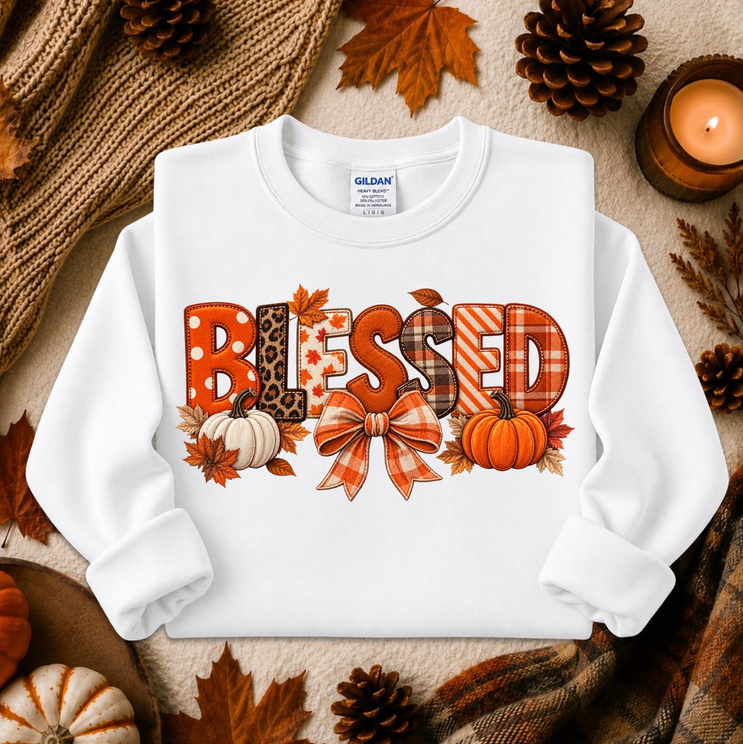 A white long-sleeve crewneck sweatshirt with a 'Blessed' autumn graphic, styled in a cozy flat-lay with fall leaves, pinecones, a candle, and a knitted blanket.