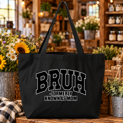 A black canvas tote bag with long handles sits on a rustic wooden table in a shop setting. The bag features the text 'BRUH -FORMERLY KNOWN AS MOM' in white varsity-style lettering.