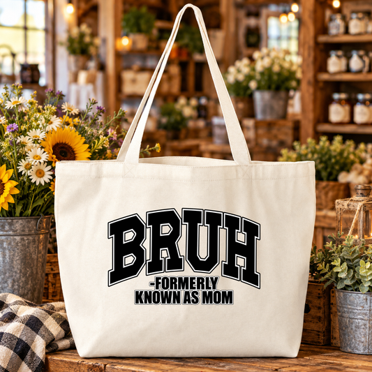 A large off-white canvas tote bag with long handles, featuring the text 'BRUH -FORMERLY KNOWN AS MOM' in black varsity-style lettering. The bag is displayed on a rustic wooden table inside a cozy, warm-toned shop setting with flowers and shelves in the background.