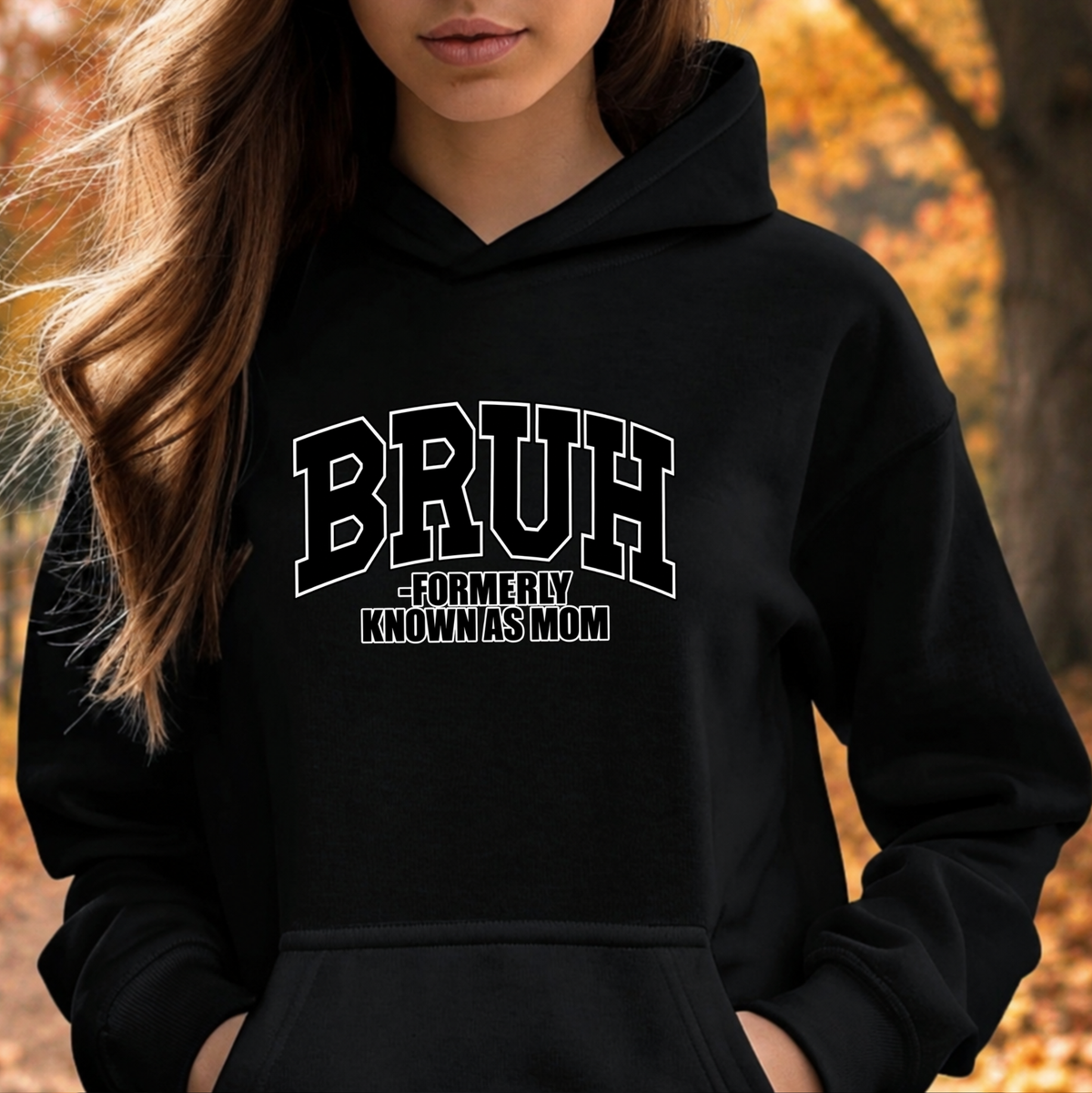A woman wearing a black pullover hoodie with a white graphic text print that reads 'BRUH -FORMERLY KNOWN AS MOM', set against an outdoor autumn background with blurred orange foliage.
